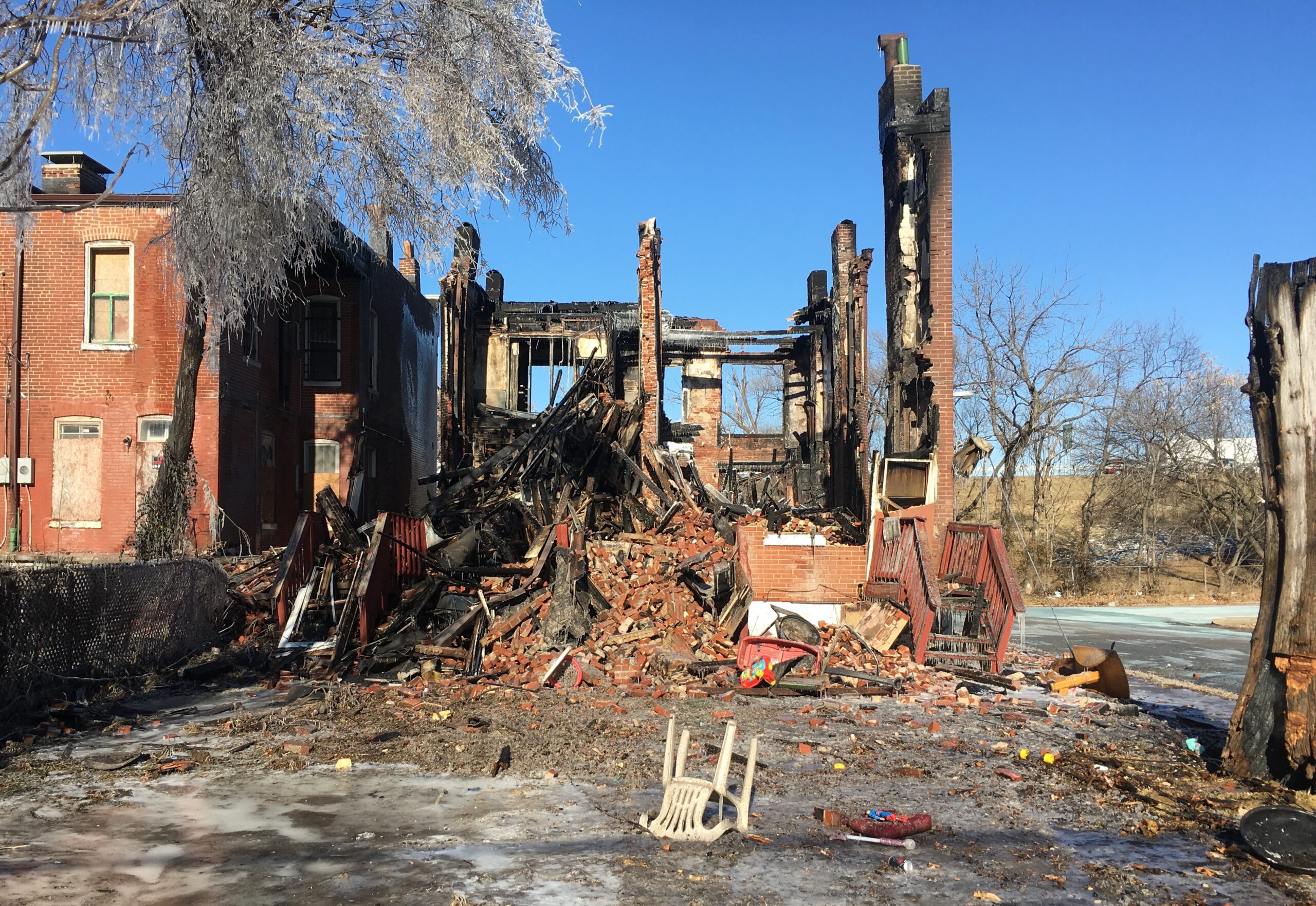 A photo of the remains of a brick quadruplex. It recently burned down, and is surrounded by ice. All that remains standing are pieces of the brick frame. A pile of bricks, rubble, and trash surround it.